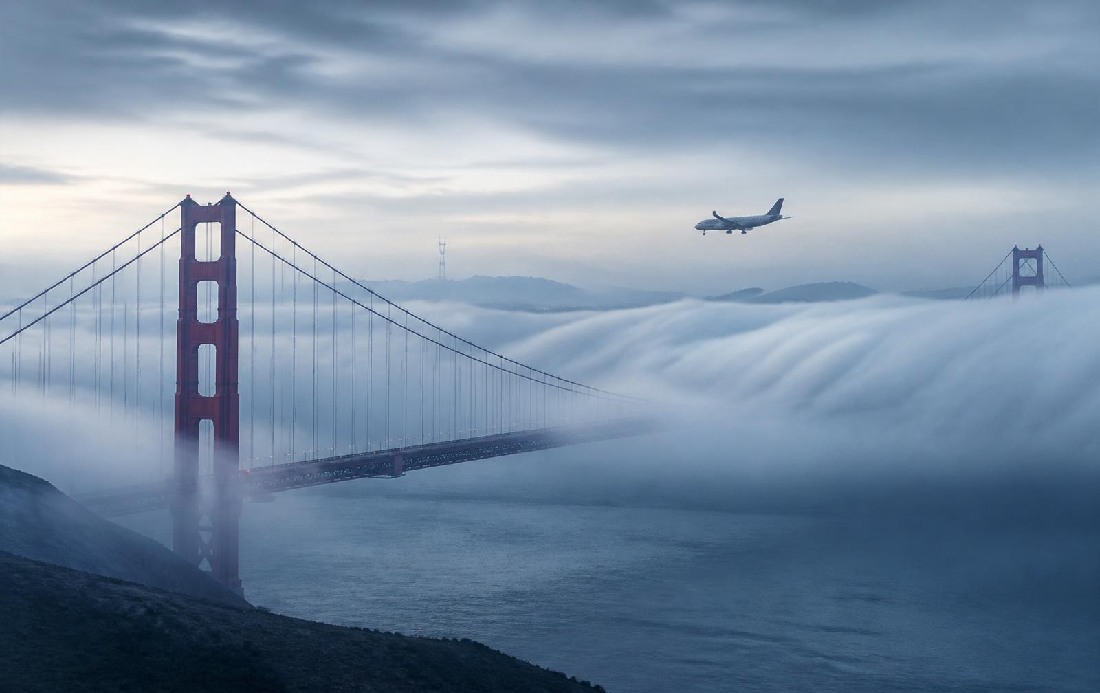 The Golden Gate Bridge cloaked in marine layer fog with an airliner on approach